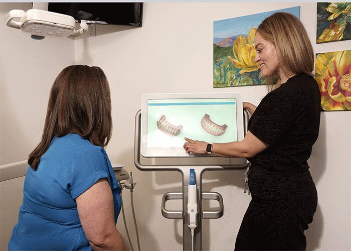 The image shows two individuals in what appears to be a dental office. One is seated, wearing a blue shirt, and the other is standing, wearing a black shirt, pointing at a screen displaying dental arches. The screen is mounted on a stand with a dental tool attached. The room has colorful paintings of flowers and landscapes on the wall.