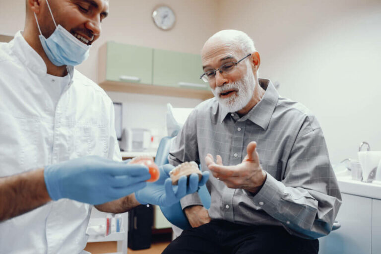 a senior aged man sits in a dental office while the dentist shows him a model of a mouth